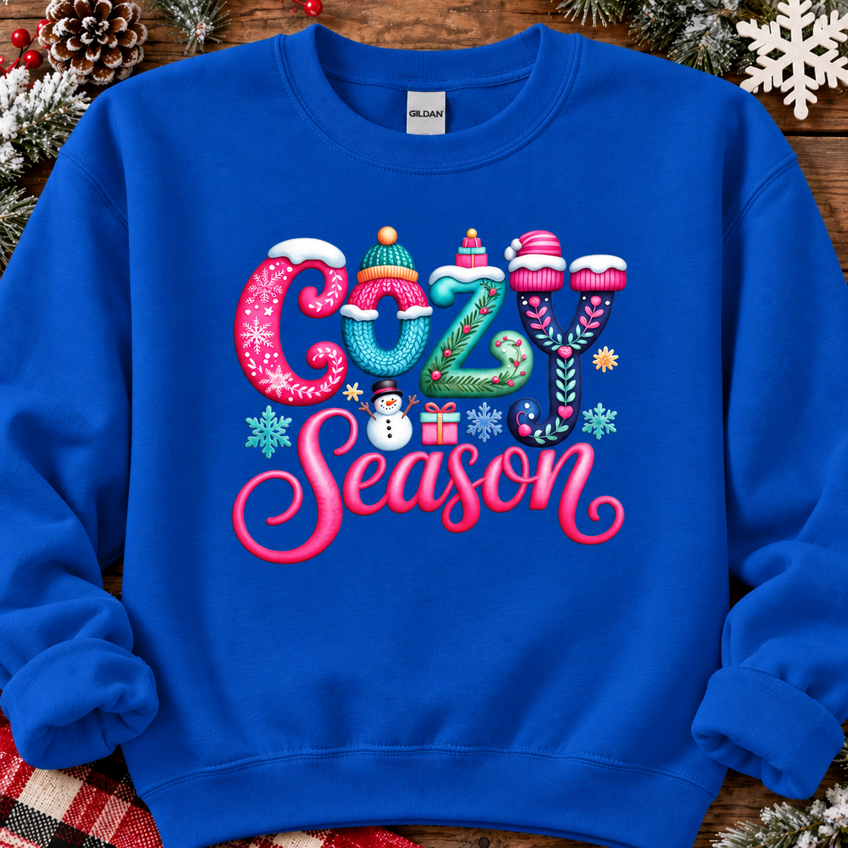 A sapphire blue crewneck sweatshirt featuring a colorful graphic that reads 'COZY Season' in knitted, winter-themed lettering. The sweatshirt is styled flat on a rustic wooden background decorated with pinecones, evergreen branches, and white snowflakes.