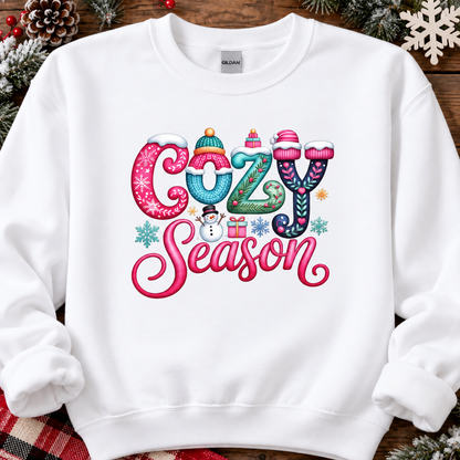 A white crewneck sweatshirt with a colorful 'Cozy Season' graphic print, styled flat on a rustic wooden background with holiday decorations like pinecones and snowflakes.