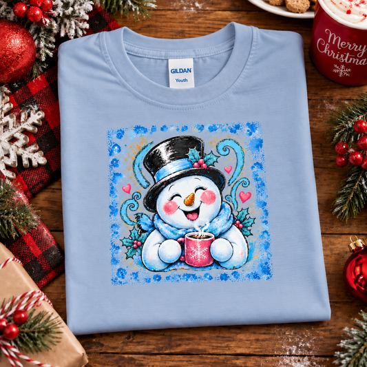 A folded light blue Gildan Youth t-shirt with a colorful Christmas snowman graphic, styled on a wooden table with holiday decorations.