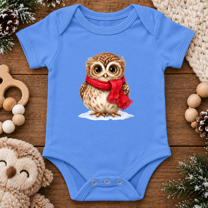 A baby bodysuit in carolina blue, featuring a graphic print of a brown owl wearing a red scarf on a snowy surface. The bodysuit is styled in a flat-lay arrangement on a rustic wooden background, surrounded by cozy props including a wooden teething ring, a plush teddy bear, pinecones, and knitted blankets.