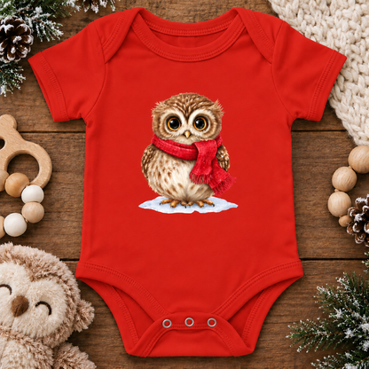 A red short-sleeve baby bodysuit laid flat on a wooden surface, featuring a printed illustration of a brown owl wearing a red scarf standing on a small patch of white snow. The bodysuit is styled with holiday props including pinecones, a plush toy, wooden beads, and a knitted blanket.
