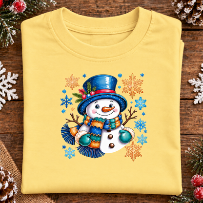 A folded butter yellow t-shirt with a colorful Christmas snowman graphic, displayed on a rustic dark wood background with pine needles and white snowflake decorations.
