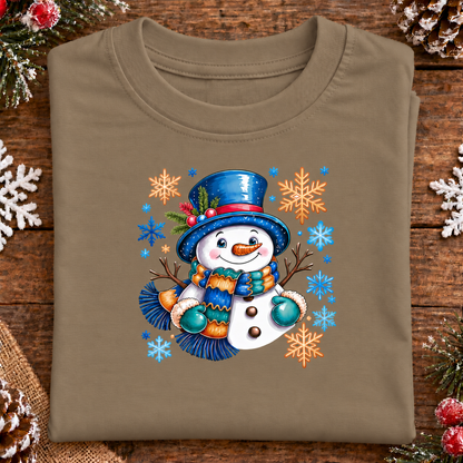 A folded coyote brown t-shirt with a colorful Christmas snowman graphic, displayed on a rustic wooden background with holiday decorations.