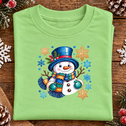 A folded keylime green crewneck t-shirt with a colorful Christmas snowman graphic, displayed on a rustic wooden surface decorated with white snowflakes and pinecones.