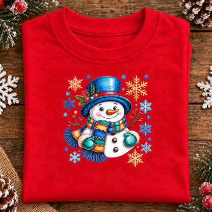 A folded red crewneck t-shirt with a colorful Christmas snowman graphic, displayed on a rustic wooden background with pine needles and white snowflakes.