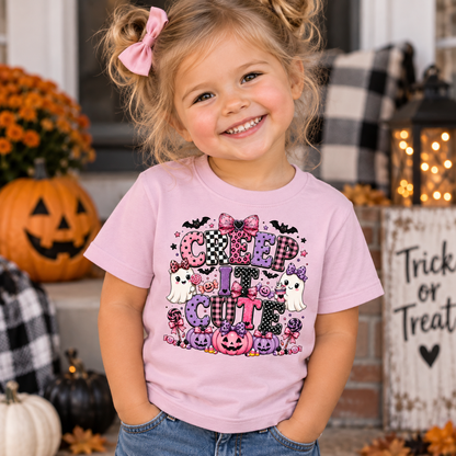 A young girl with blonde hair in a pink bow tie is smiling and wearing a light pink graphic t-shirt that says 'CUTE' in a Halloween-themed font with pumpkins, ghosts, and bats. She is standing outdoors in a decorated fall setting with pumpkins and a 'Trick or Treat' sign.