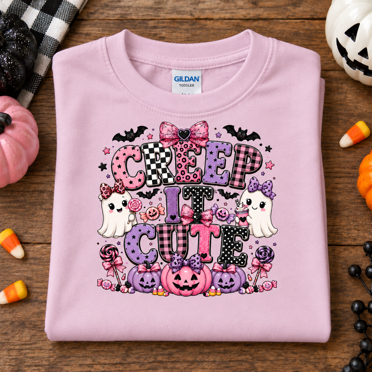 A folded light pink Gildan t-shirt with a colorful 'CREEP IT CUTE' Halloween graphic, displayed on a rustic wooden table surrounded by seasonal props like candy corn, pumpkins, and a plaid blanket.