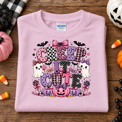 A folded light pink Gildan t-shirt with a colorful 'CREEP IT CUTE' Halloween graphic, displayed on a rustic wooden table surrounded by seasonal props like candy corn, pumpkins, and a plaid blanket.