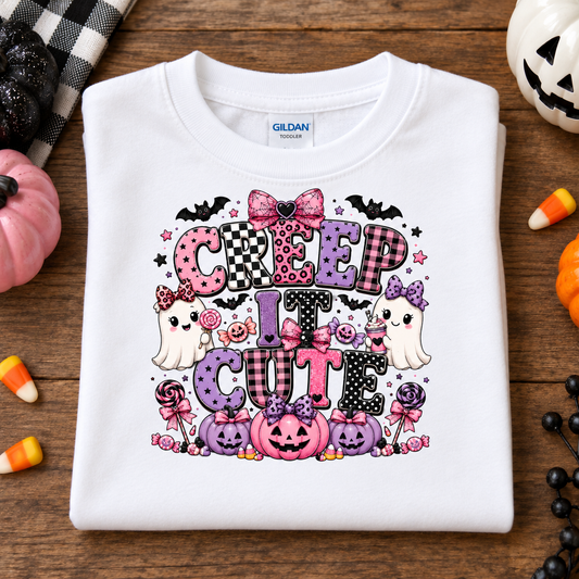 A folded white Gildan T-shirt with a colorful Halloween graphic reading 'CREEP IT CUTE' in pink, purple, black, and white tones, surrounded by cartoon ghosts, pumpkins, bats, and candy, displayed on a wooden surface with seasonal props.