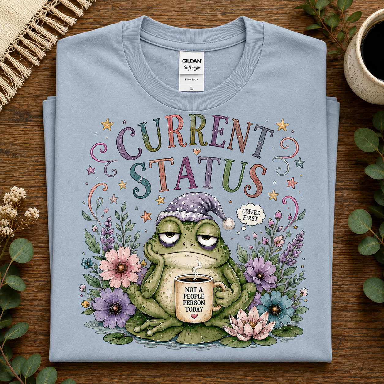 A folded light blue Gildan Softstyle t-shirt is displayed on a wooden surface with decorative plants and a cup. The shirt features a colorful graphic of a sleeping frog wearing a beanie and holding a mug that reads 'COFFEE FIRST' and 'NOT A PEOPLE'S PERSON TODAY'.