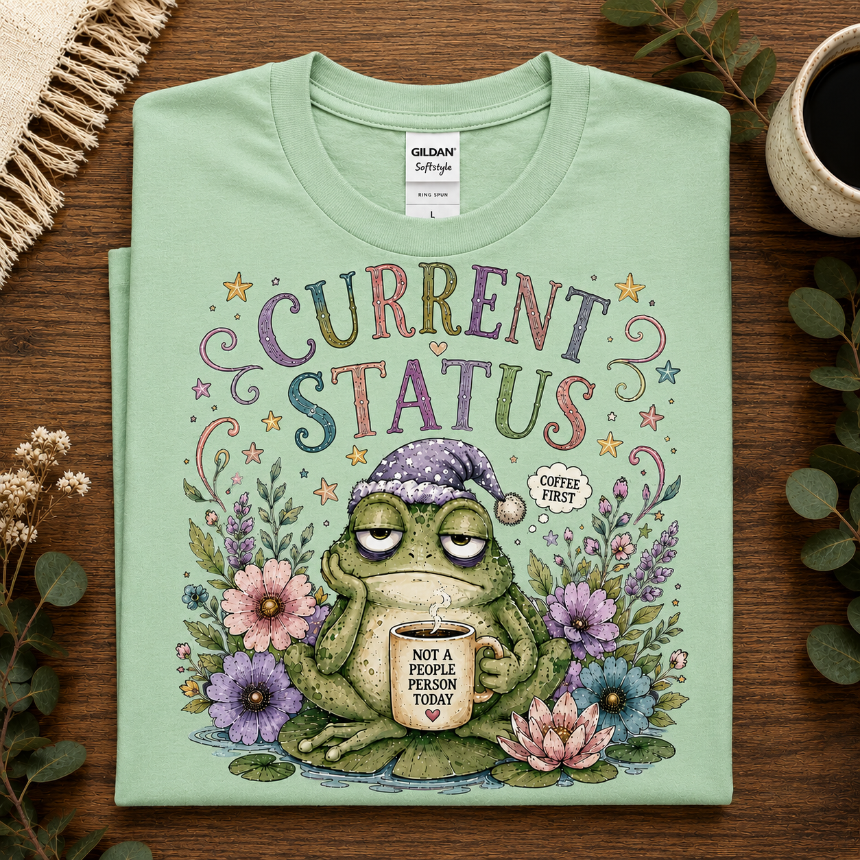 A folded mint green Gildan Softstyle t-shirt is displayed flat on a dark wooden table. The shirt features a colorful graphic of a tired green frog wearing a purple winter hat and holding a mug that says 'NOT A PEOPLE PERSON TODAY'.