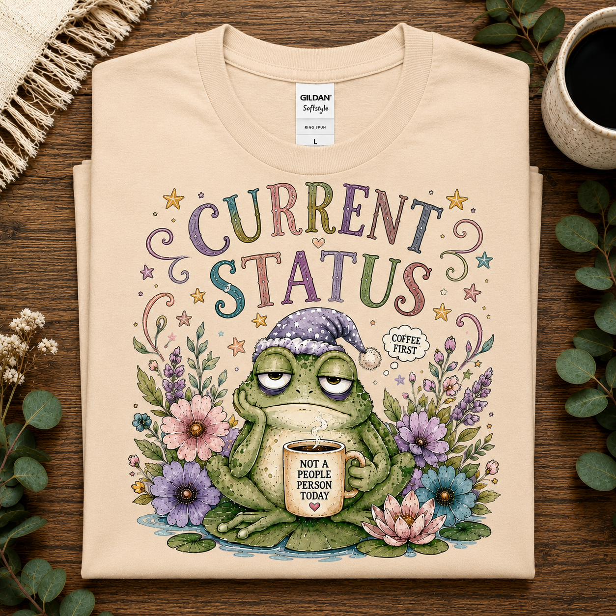 A folded natural colored t-shirt with a colorful graphic print of a sleeping green frog holding a coffee mug surrounded by flowers and the text 'CURRENT STATUS COFFEE FIRST'. The t-shirt is displayed on a wooden surface with decorative props including a mug of coffee, eucalyptus leaves, and a fringed textile.