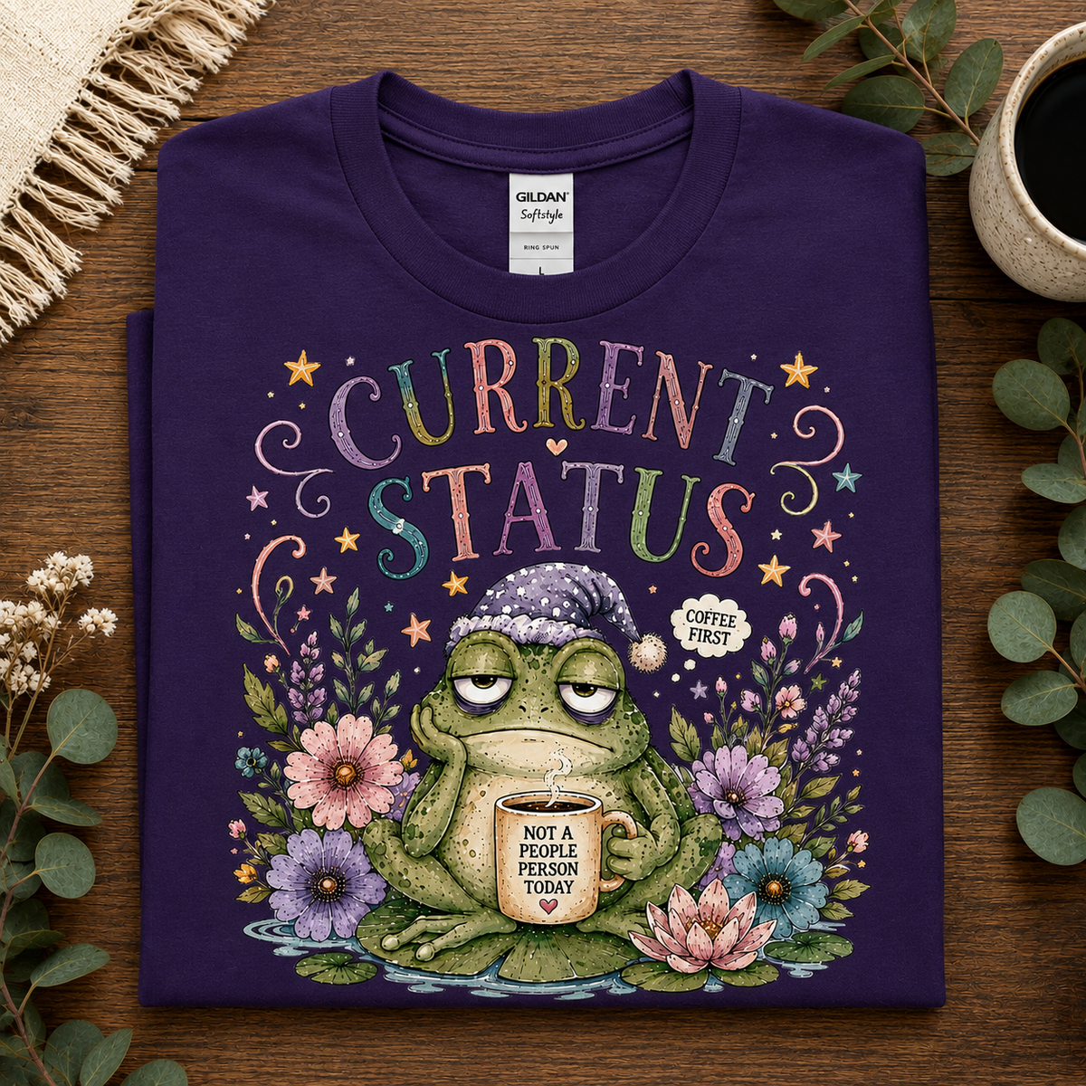 A folded dark purple Gildan Softstyle t-shirt with a colorful graphic of a green frog wearing a beanie and holding a mug that says 'NOT A PEOPLE PERSON TODAY'. The shirt is displayed on a wooden table with decorative eucalyptus leaves, a cup of coffee, and a woven textile.