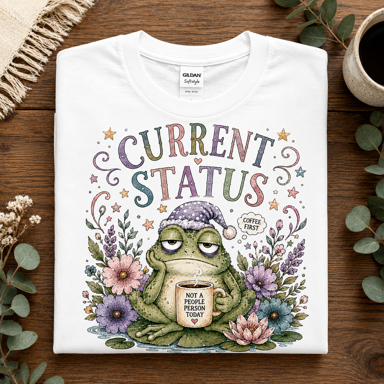 A folded white Gildan Softstyle T-shirt with a colorful graphic of a sleeping green frog holding a coffee mug under the words 'CURRENT STATUS COFFEE FIRST', styled on a wooden surface with decorative greenery and a cup of coffee.
