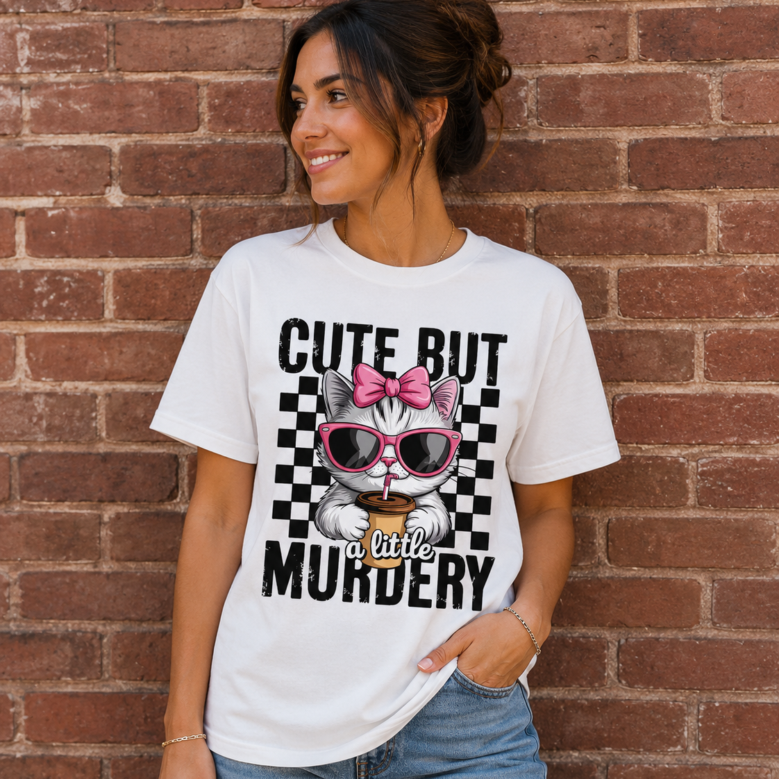 Woman wearing a white graphic t-shirt with a cute kitten wearing pink sunglasses and the phrase “Cute But a Little Murdery” standing in front of a brick wall.