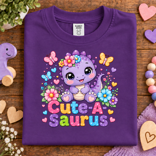 A folded purple t-shirt with a colorful 'Cute-A Saurus' graphic, displayed on a wooden surface surrounded by decorative props like wooden hearts, beads, and a small plant.