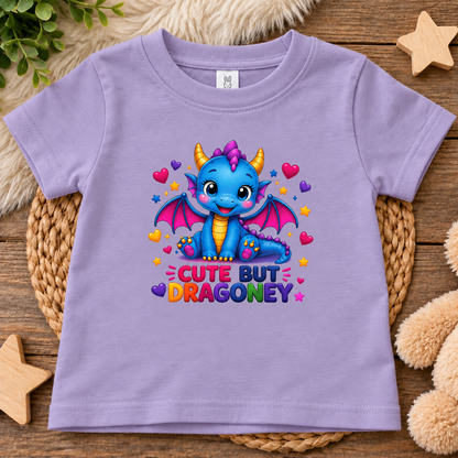 A lavender short-sleeve kids' t-shirt with a colorful cartoon dragon graphic and the text 'CUTE BUT DRAGONEY' printed on the front. The t-shirt is displayed flat on a woven placemat over a wooden surface with decorative plush toys and a star.