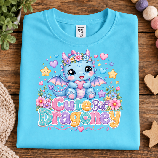 A folded aqua blue t-shirt with a colorful graphic of a cute blue dragon and the text 'Cute But Dragony' in pastel letters, displayed on a wooden surface with decorative props.