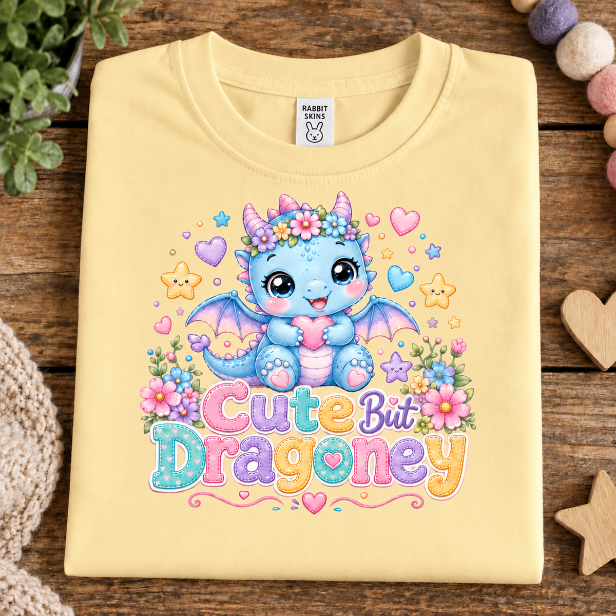A pale butter yellow folded T-shirt laid flat on a rustic wooden surface, featuring a colorful cartoon dragon graphic with flowers and the text 'Cute But Dragony.' A Rabbit Snaps neck label is visible.