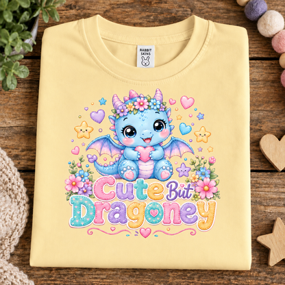 A pale butter yellow folded T-shirt laid flat on a rustic wooden surface, featuring a colorful cartoon dragon graphic with flowers and the text 'Cute But Dragony.' A Rabbit Snaps neck label is visible.