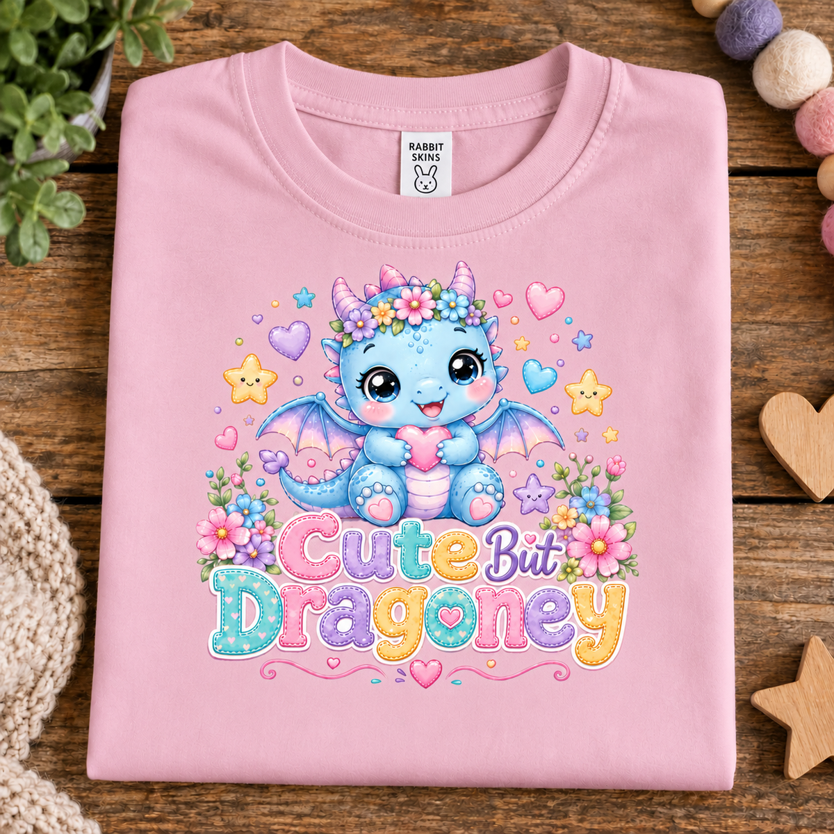 A folded pink t-shirt with a colorful graphic of a cute blue dragon and the text 'Cute But Dragoney'. The t-shirt is displayed on a rustic wooden background with decorative props like a knit blanket, wooden hearts, and a plant.