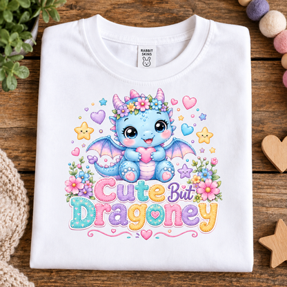 A folded white t-shirt with a colorful graphic of a cute blue dragon and the text 'Cute But Dragoney' on a rustic wooden background.