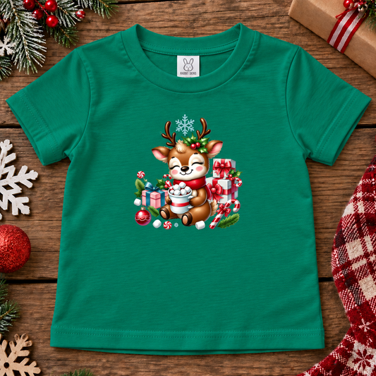 A toddler's short-sleeve green t-shirt with a festive Christmas graphic of a reindeer holding a mug of hot cocoa, surrounded by gifts, candy canes, and snowballs. The shirt is displayed flat on a rustic wooden background decorated with holiday ornaments, pine branches, and a plaid scarf.