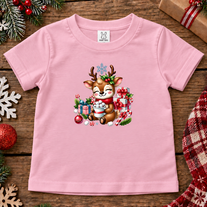 A light pink short-sleeve baby t-shirt with a colorful Christmas cartoon deer graphic, laid flat on a rustic wooden background decorated with holiday ornaments, pine branches, and a plaid blanket.