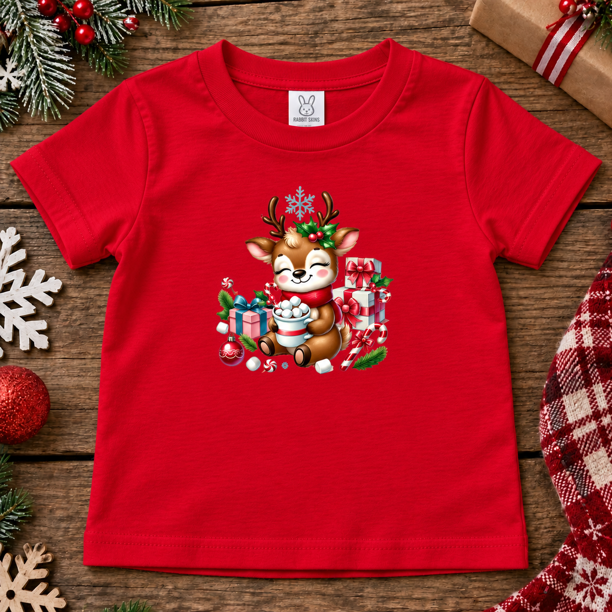 A red short-sleeve baby t-shirt with a festive Christmas graphic of a smiling reindeer surrounded by gifts, displayed on a rustic wooden background with holiday decorations.