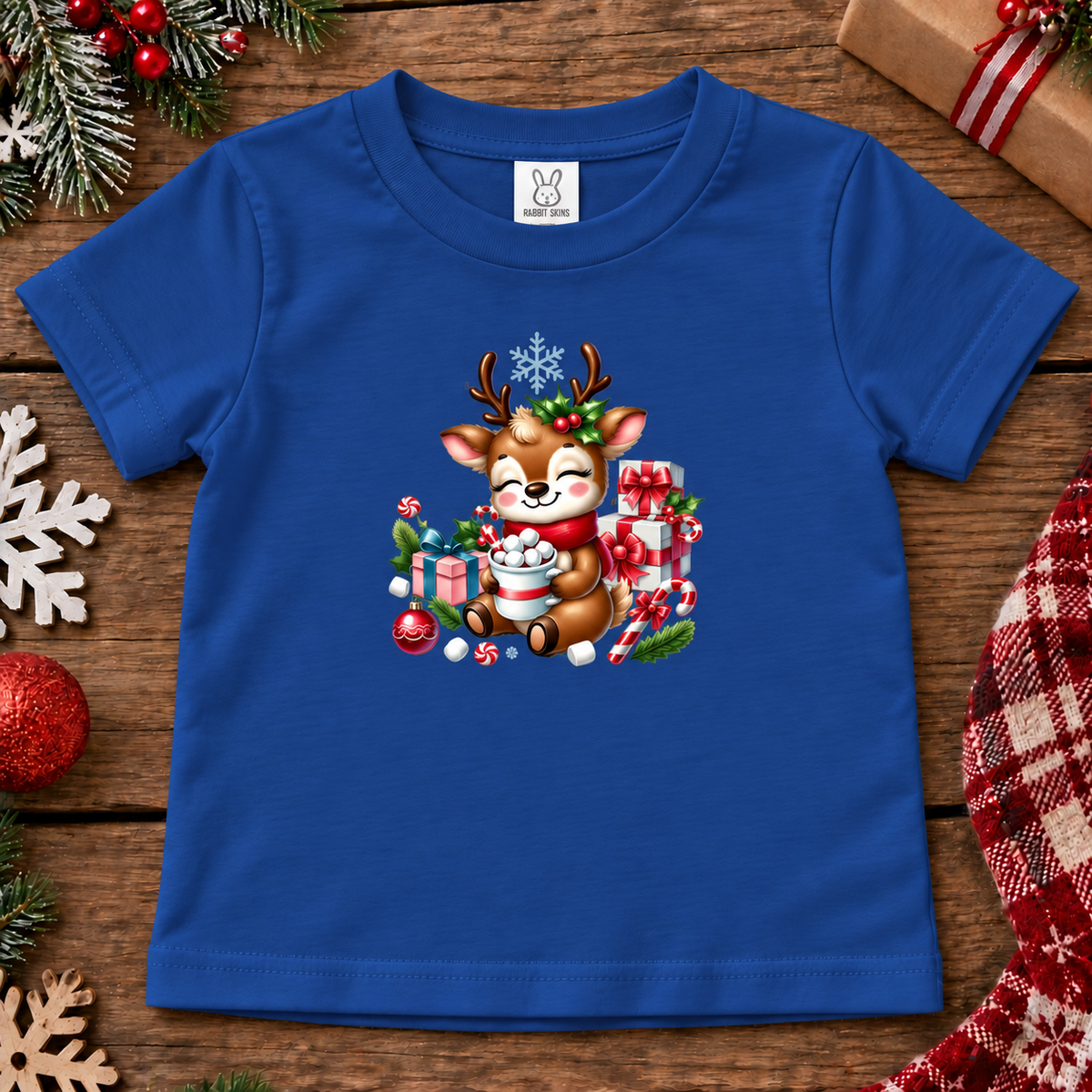 A royal blue short-sleeve baby t-shirt with a colorful Christmas cartoon deer graphic, laid flat on a rustic wooden surface surrounded by holiday decorations.