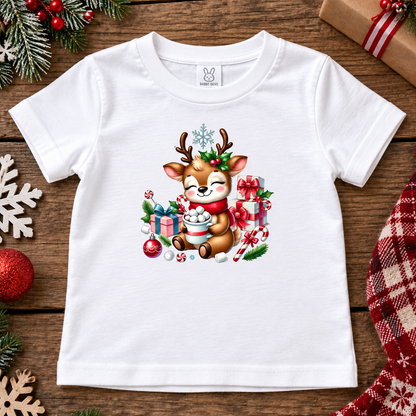 A white short-sleeve baby t-shirt with a colorful Christmas-themed graphic of a cute cartoon reindeer holding hot cocoa, surrounded by gifts, candy canes, and ornaments, laid flat on a rustic wooden background with holiday decorations.