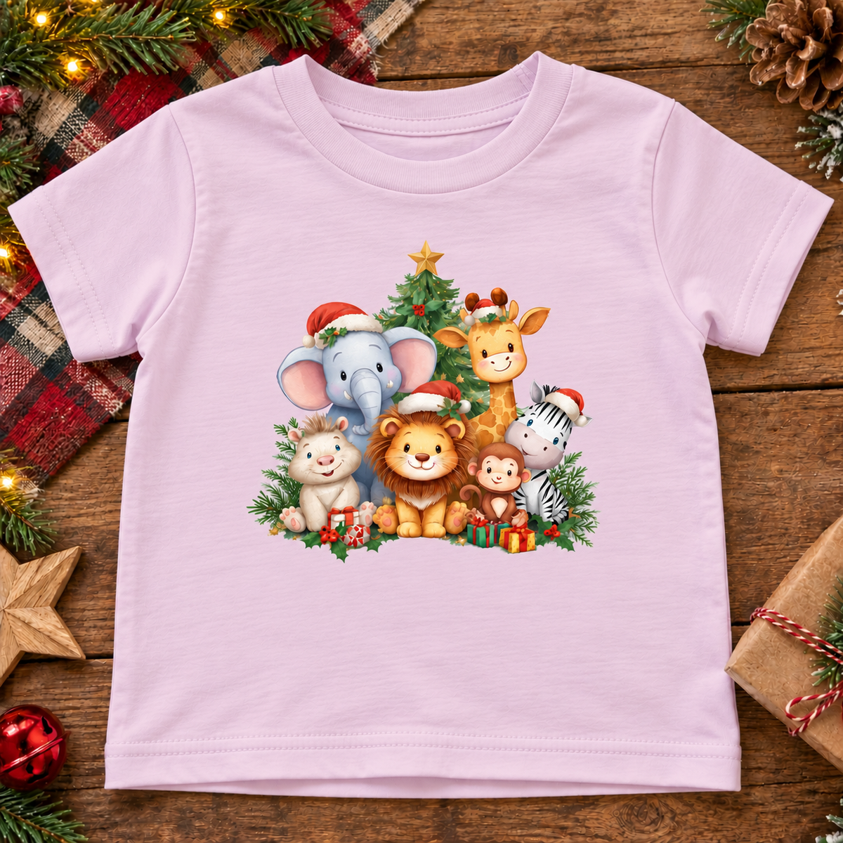 A bllerina pink short-sleeve baby t-shirt with a colorful Christmas animal graphic, laid flat on a rustic wooden surface surrounded by holiday decorations like pine branches, a plaid blanket, and wrapped gifts.