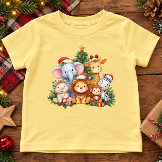 A butter yellow toddler t-shirt featuring a colorful Christmas safari animal graphic, laid flat on a rustic wooden surface decorated with pine branches, a plaid scarf, and wrapped gifts.