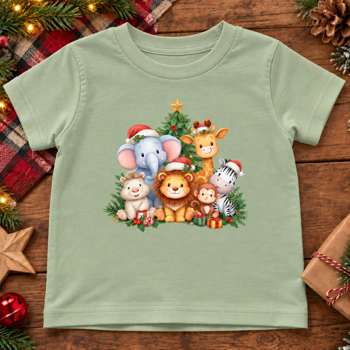 A sage green short-sleeve baby t-shirt with a colorful cartoon animal Christmas graphic, displayed flat on a rustic wooden background with holiday decorations.