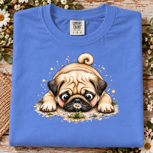 A folded mystic blue Comfort Colors t-shirt featuring a cute graphic of a pug puppy holding a daisy, displayed on a rustic wooden surface surrounded by white flowers.