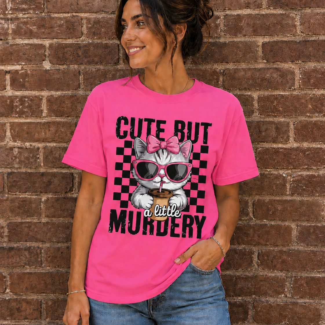 Woman wearing a Neon Pink graphic t-shirt with a cute kitten wearing pink sunglasses and the phrase “Cute But a Little Murdery” standing in front of a brick wall.