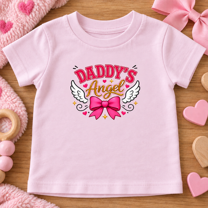 A ballerina pink short-sleeve baby t-shirt with a 'Daddy's Angel' graphic on the chest, styled in a flat-lay arrangement on a wooden surface with pink toys.