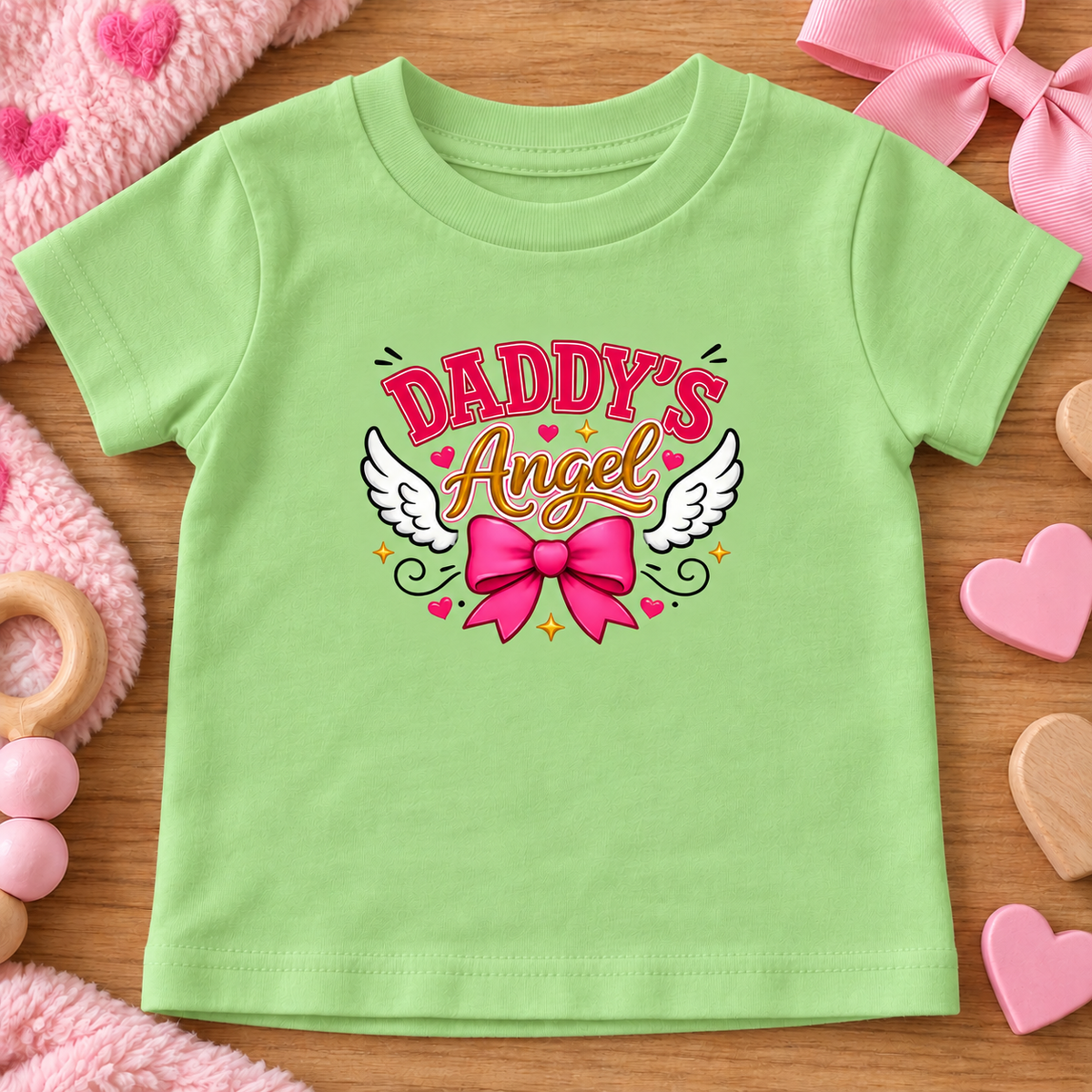 A keylime green baby t-shirt with a 'Daddy's Angel' graphic featuring white wings, a pink bow, and hearts, laid flat on a wooden surface with pink baby accessories.