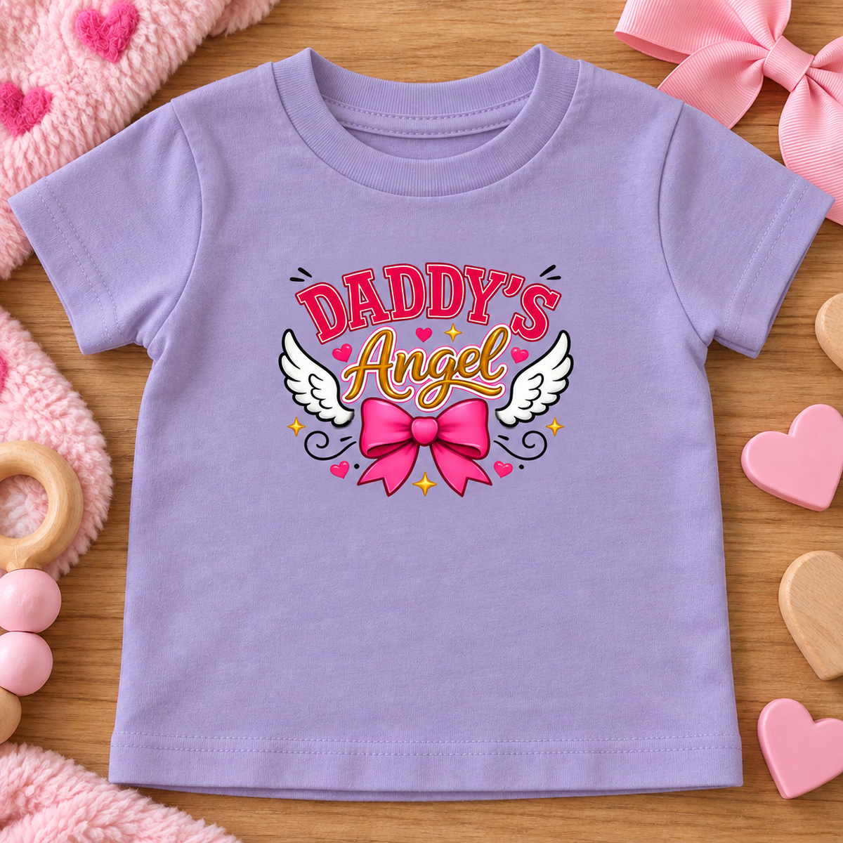 A lavender baby t-shirt with a 'Daddy's Angel' graphic featuring pink and gold lettering, white angel wings, and a pink bow, laid flat on a wooden surface with pink baby accessories.