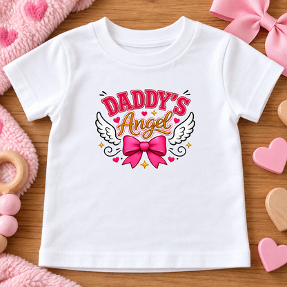 A white short-sleeve baby t-shirt with a 'Daddy's Angel' graphic print, laid flat on a wooden surface surrounded by pink baby toys.
