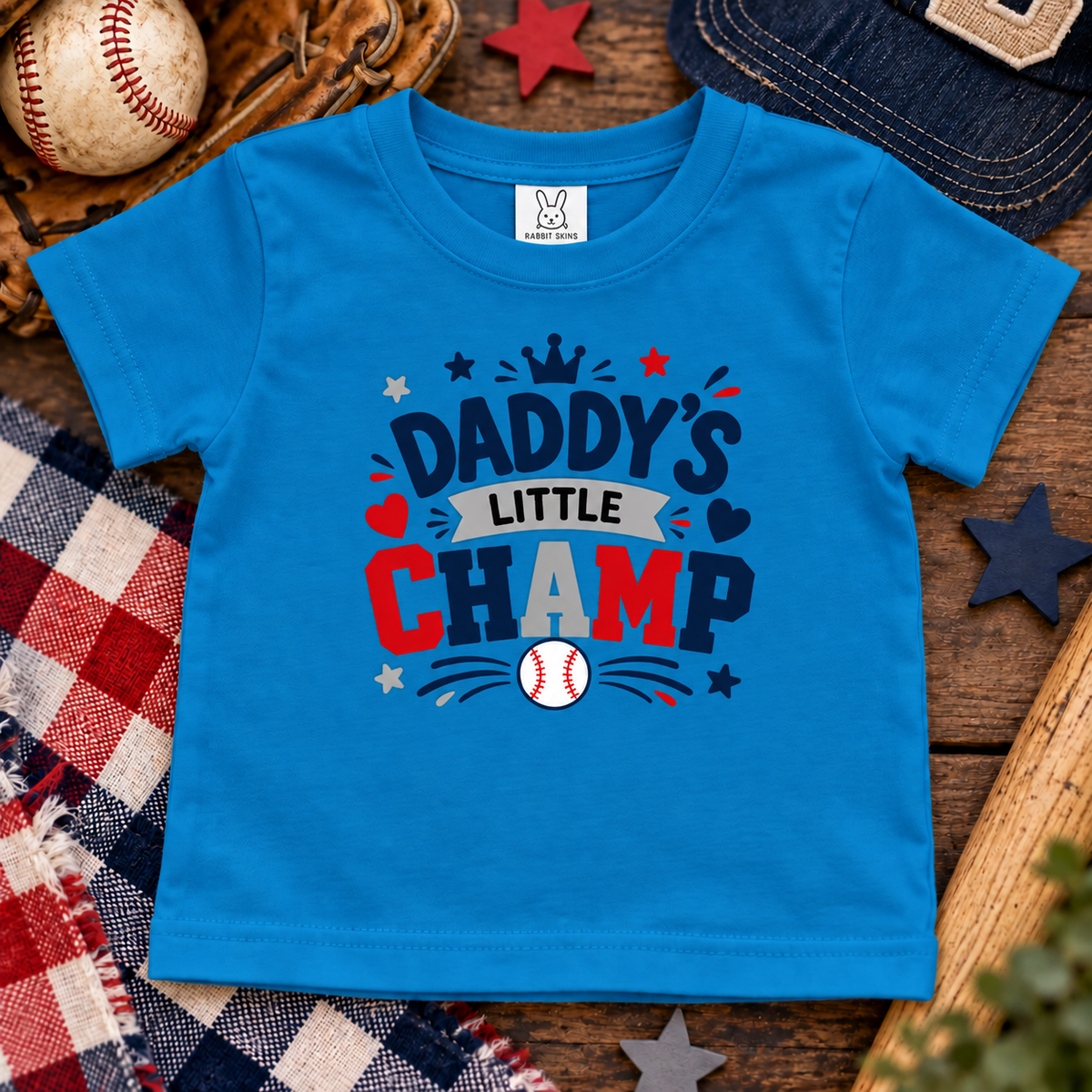 A bright cobalt short-sleeve t-shirt laid flat on a rustic wooden surface with baseball-themed props, including a baseball, glove, cap, stars, and plaid fabric. The shirt features a large front graphic reading “Daddy’s Little Champ” in navy, red, white, and gray with small baseball and star details.