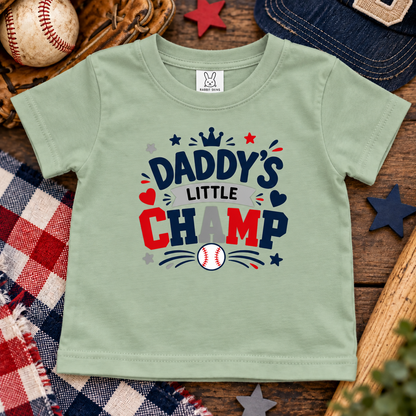 A sage green short-sleeve t-shirt for toddlers, featuring a 'Daddy's Little Champ' baseball graphic in navy, red, gray, and white. The shirt is displayed in a flat-lay arrangement with baseball-themed props including a baseball, a wooden glove, a denim cap, and a plaid blanket on a rustic wooden surface.