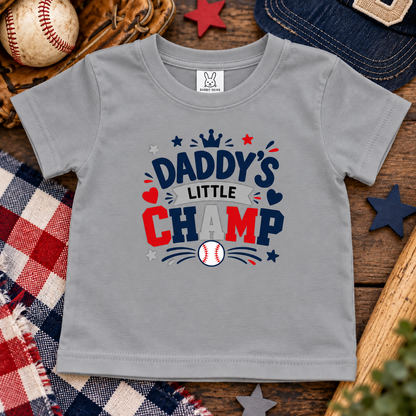 A titanium gray short-sleeve t-shirt with a 'Daddy's Little Champ' baseball graphic, displayed in a flat-lay arrangement with a baseball, glove, denim cap, and plaid blanket.