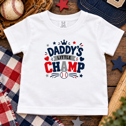 A white short-sleeve baby t-shirt with a 'Daddy's Little Champ' baseball graphic, styled in a flat-lay arrangement with a baseball, wooden glove, denim cap, and plaid blanket.