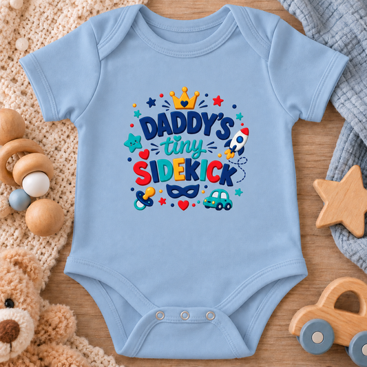 A light blue baby bodysuit laid flat on a wooden surface, surrounded by soft toys and knitted blankets. The front has a colorful graphic that reads “Daddy’s tiny sidekick” with a crown, stars, hearts, a rocket, and toy illustrations.
