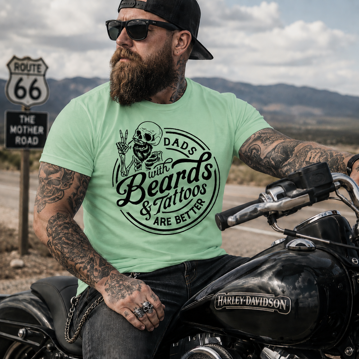Man with tattoos and beard sitting on a motorcycle wearing a mint green colored t-shirt with a graphic design.