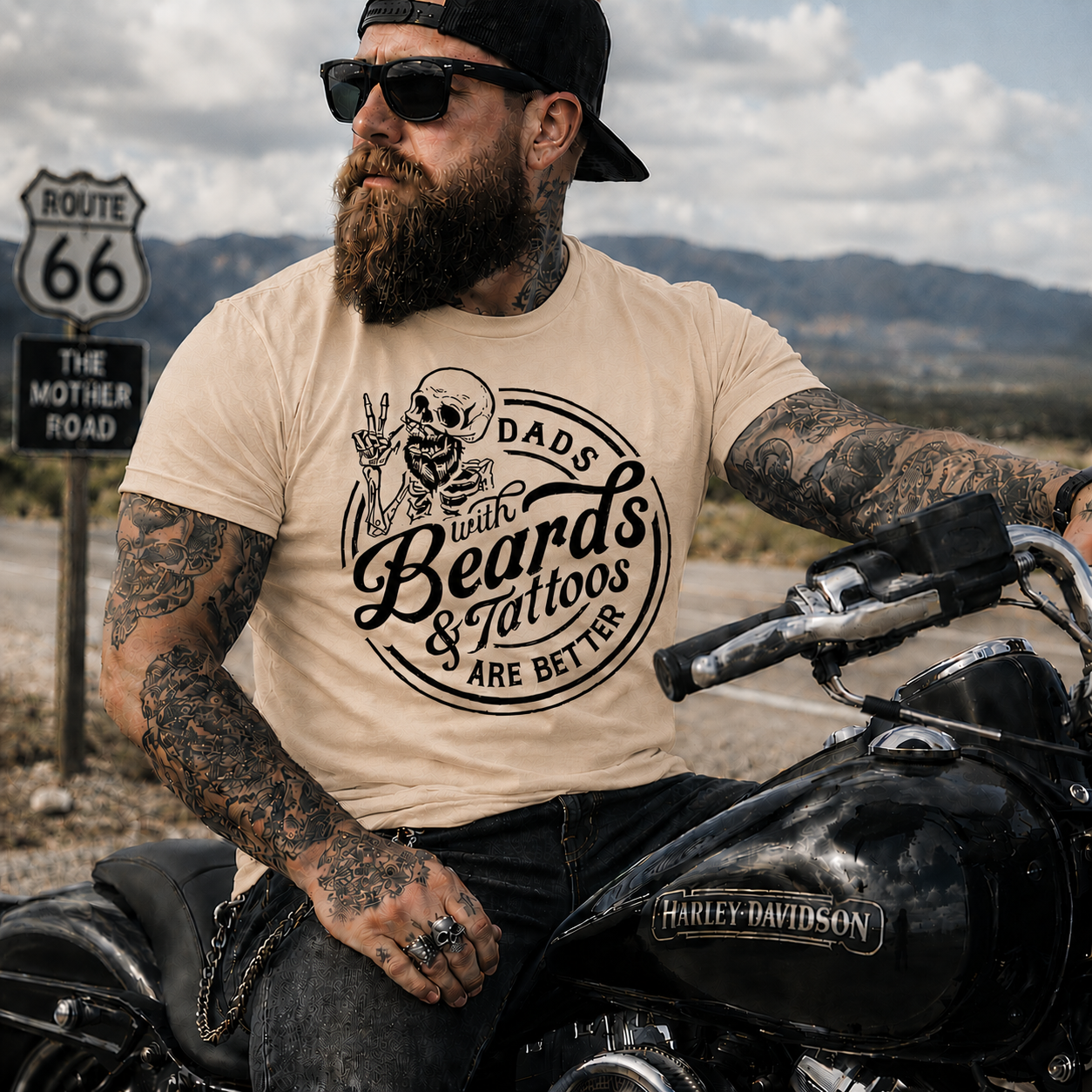 Man with tattoos and beard sitting on a motorcycle wearing a natural colored t-shirt with a graphic design.