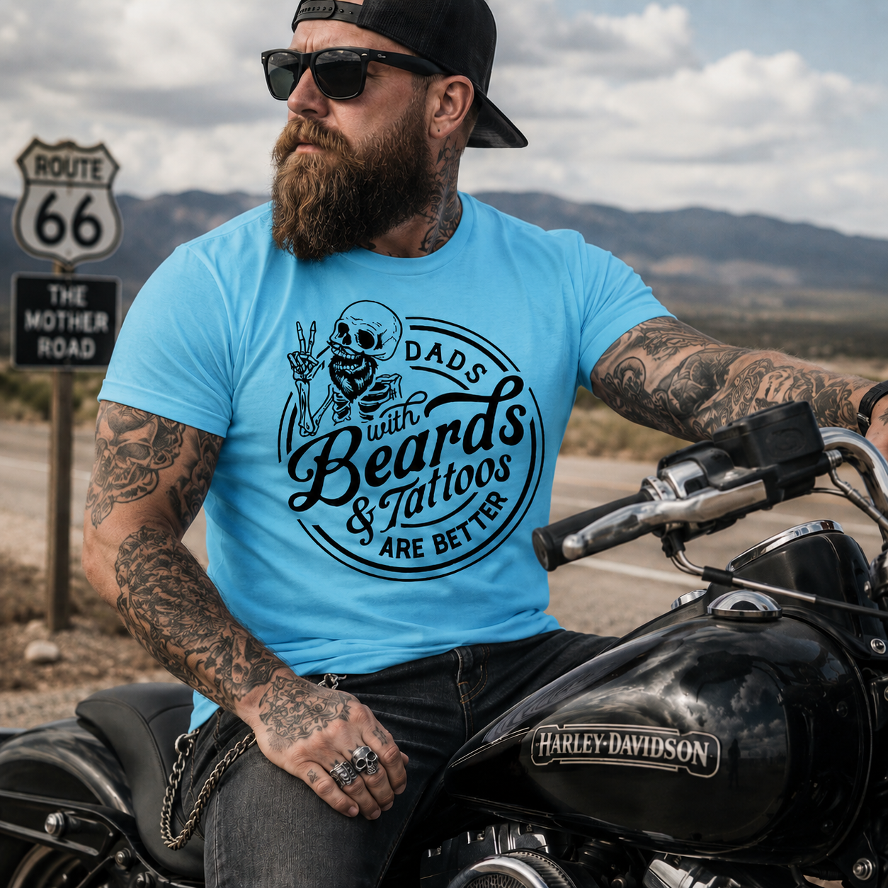 Man with tattoos and beard sitting on a motorcycle wearing a sky blue colored  t-shirt with a graphic design.