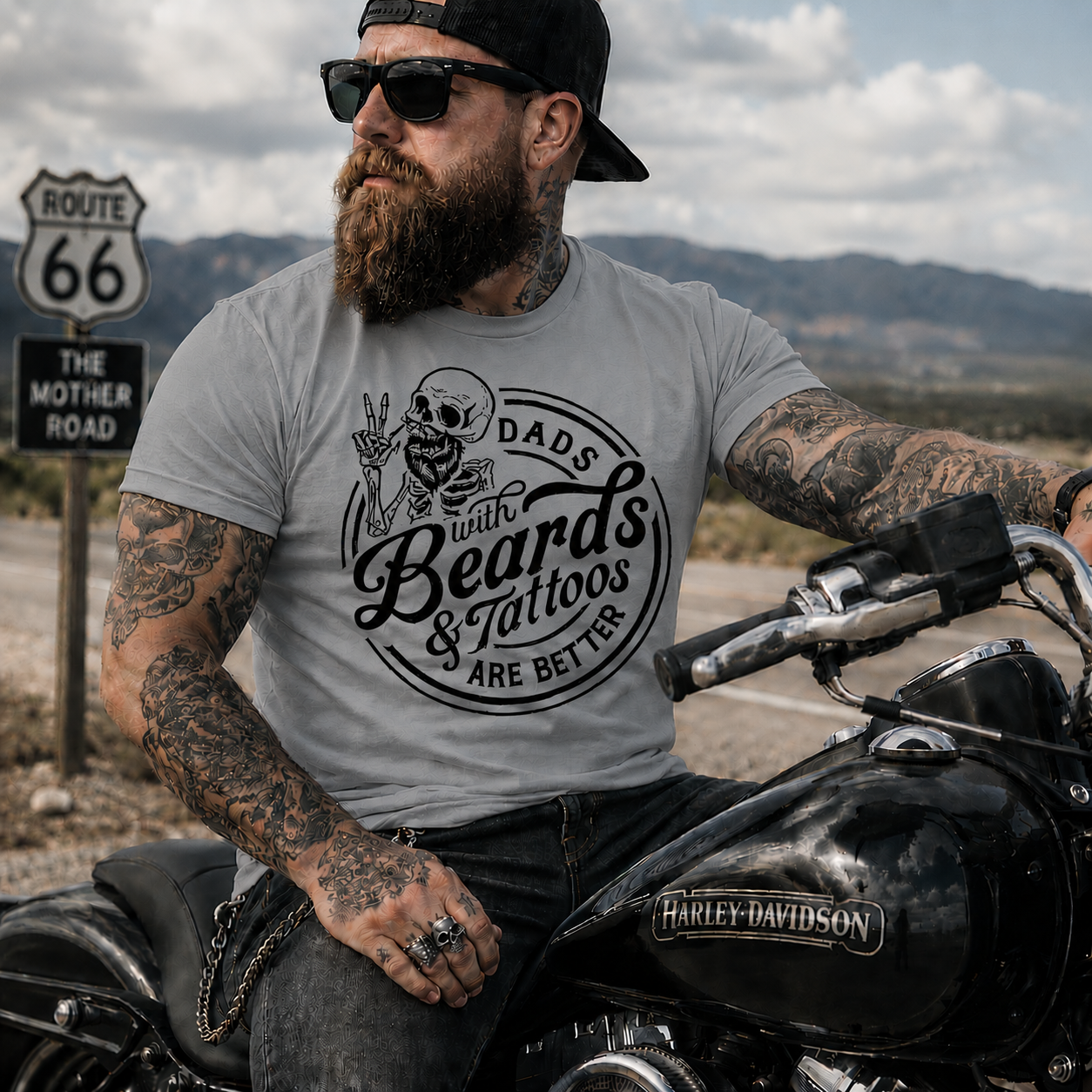Man with tattoos and beard sitting on a motorcycle wearing a sport gray colored t-shirt with a graphic design.