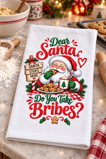 A white kitchen tea towel with a festive Christmas design featuring Santa Claus, cookies, holly, and the text 'Dear Santa Do You Take Bribes?' laid on a marble countertop with baking props in the background.